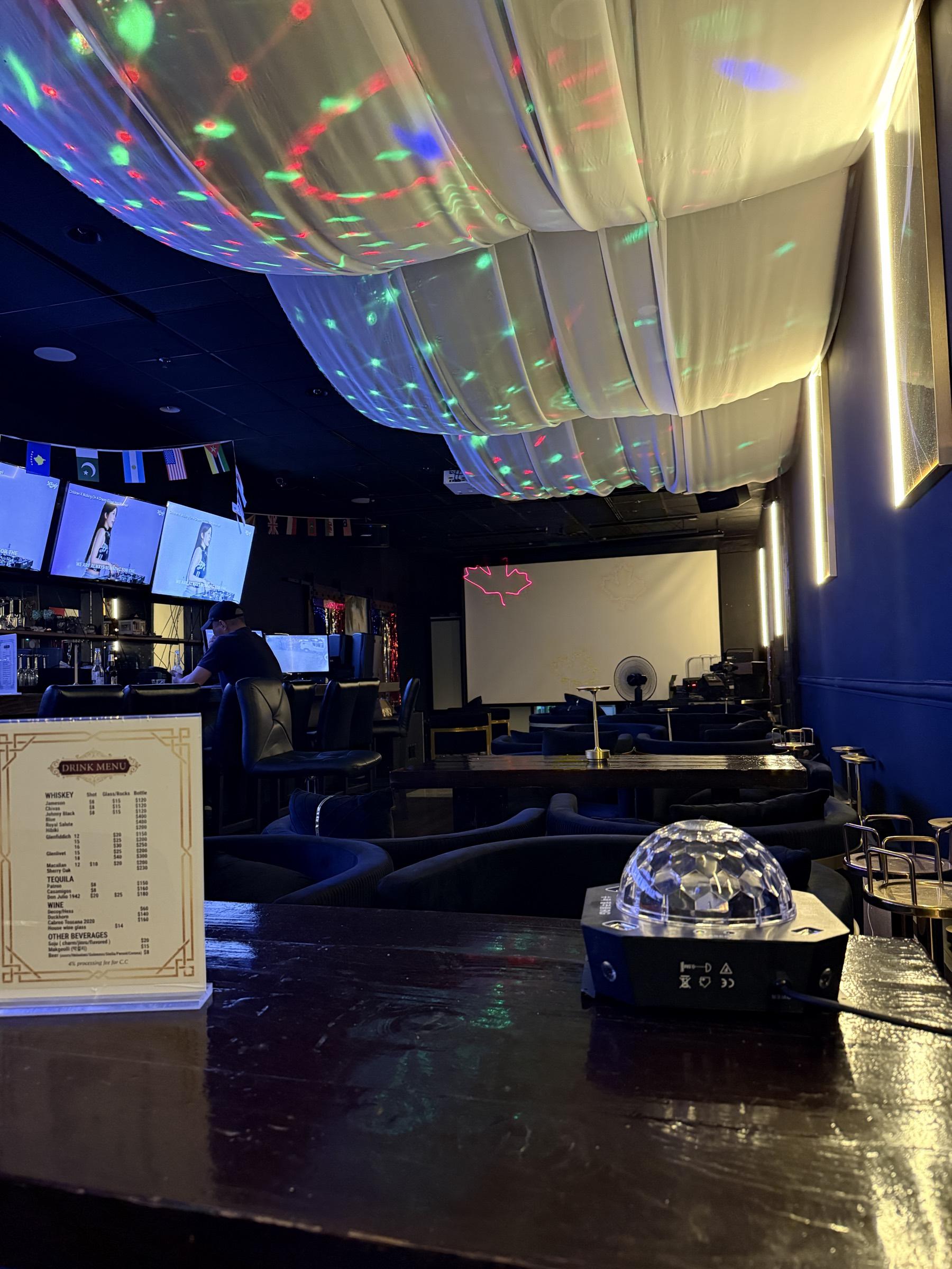 The lounge seen across the bar: coloured light scattered over the draped ceiling, screens and flags along the wall, and rows of velvet seating running back to the projection screen.