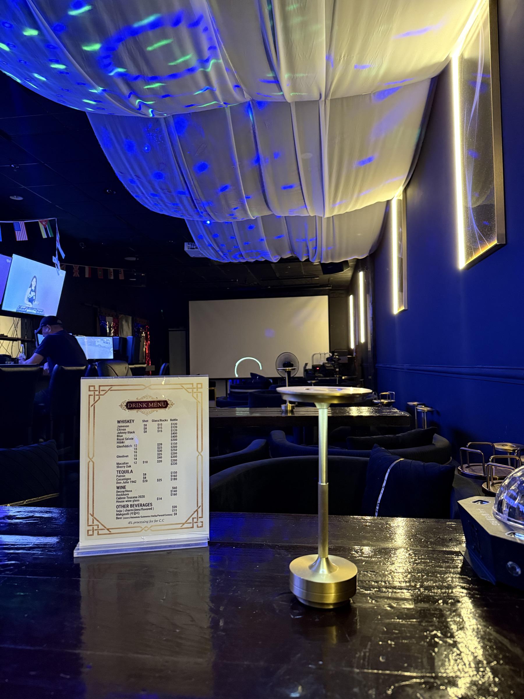 The bar top with the printed drink menu standing on it, a brass lamp beside it, and coloured light scattered across the ivory draped ceiling above the navy room.