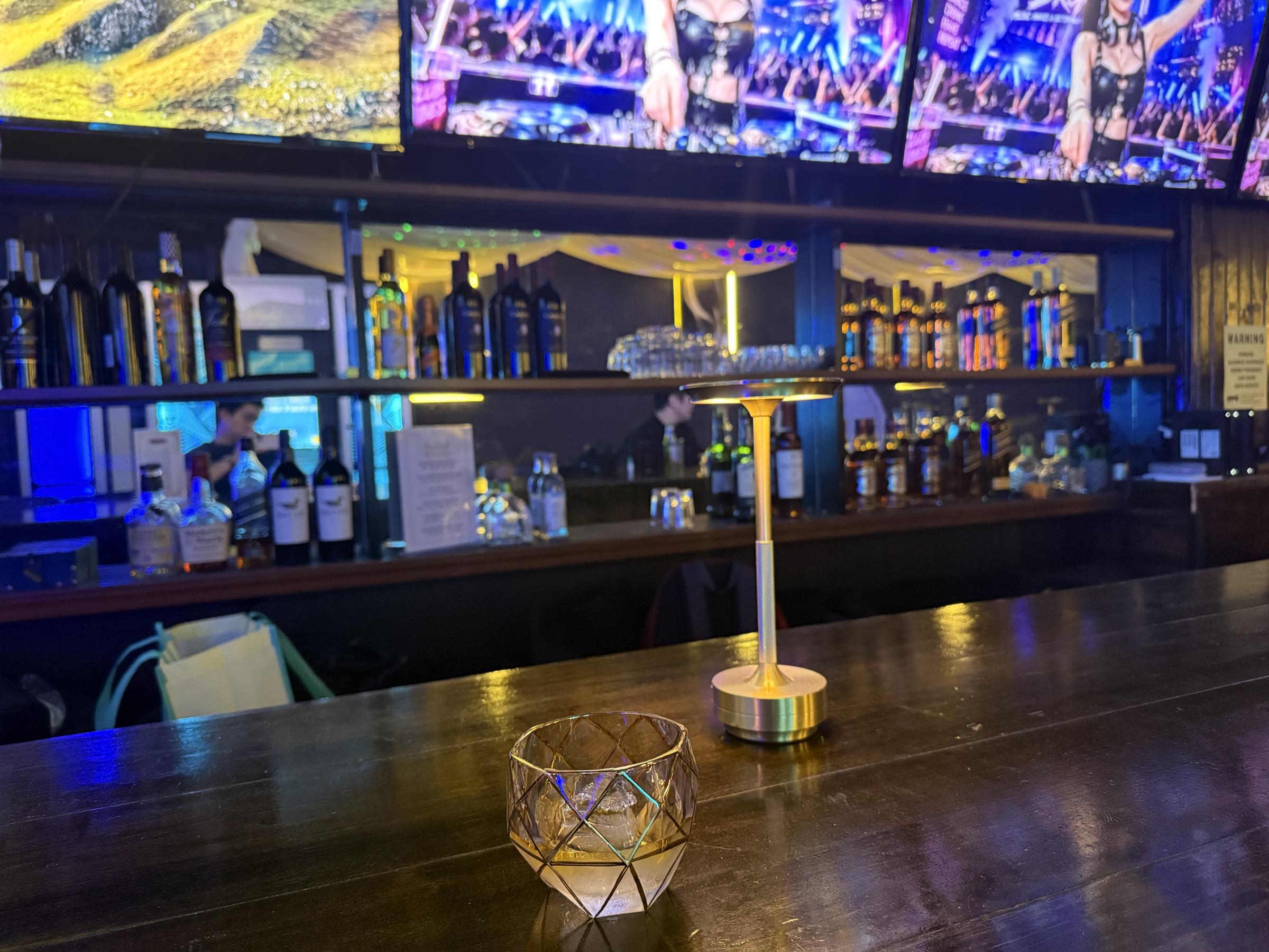 A faceted rocks glass with a gold rim on the dark wood bar top, beside a brass cordless table lamp.
