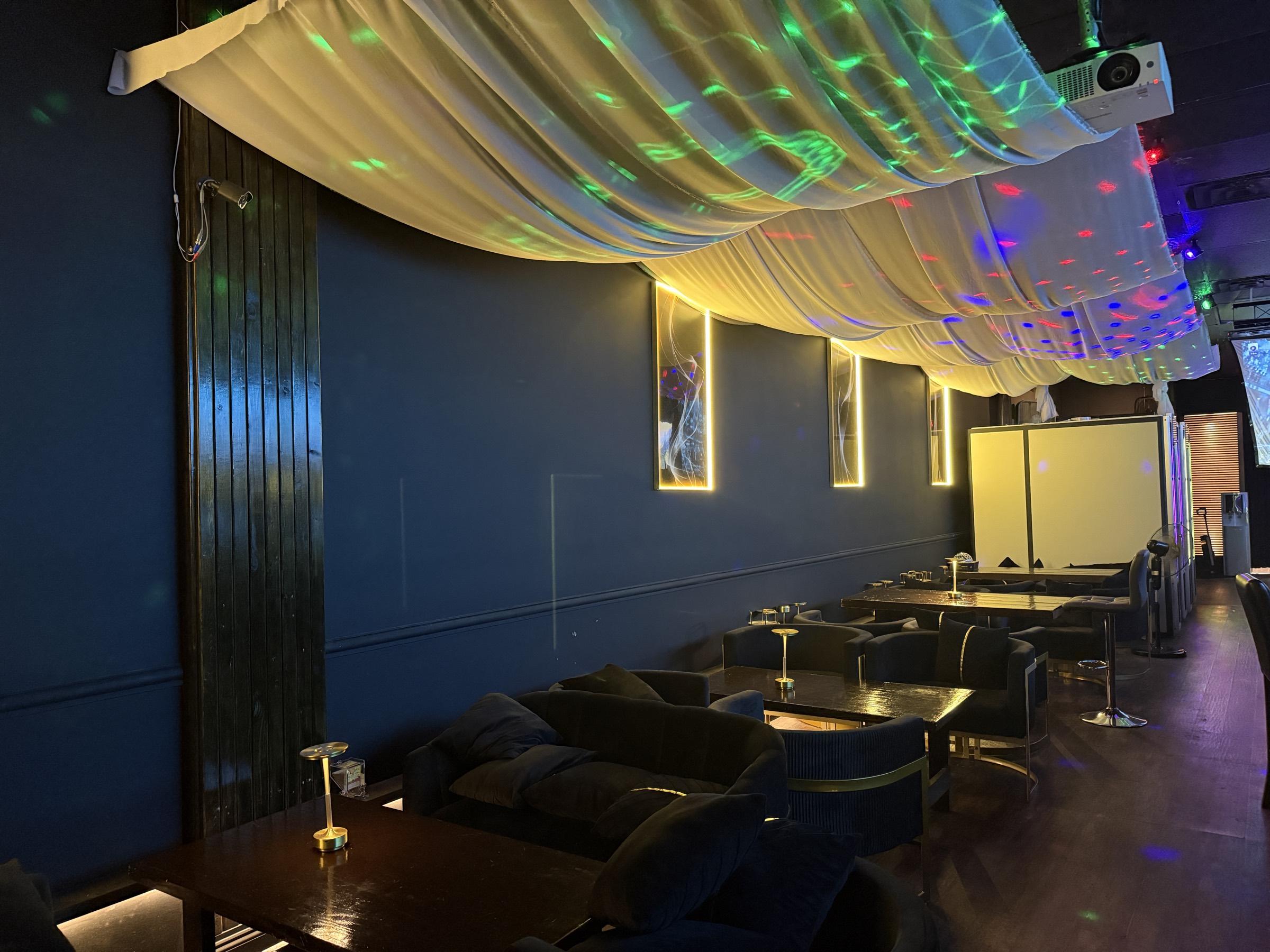 The lounge seen from the far end, with a ceiling projector overhead and coloured lights scattered across the draped ceiling.