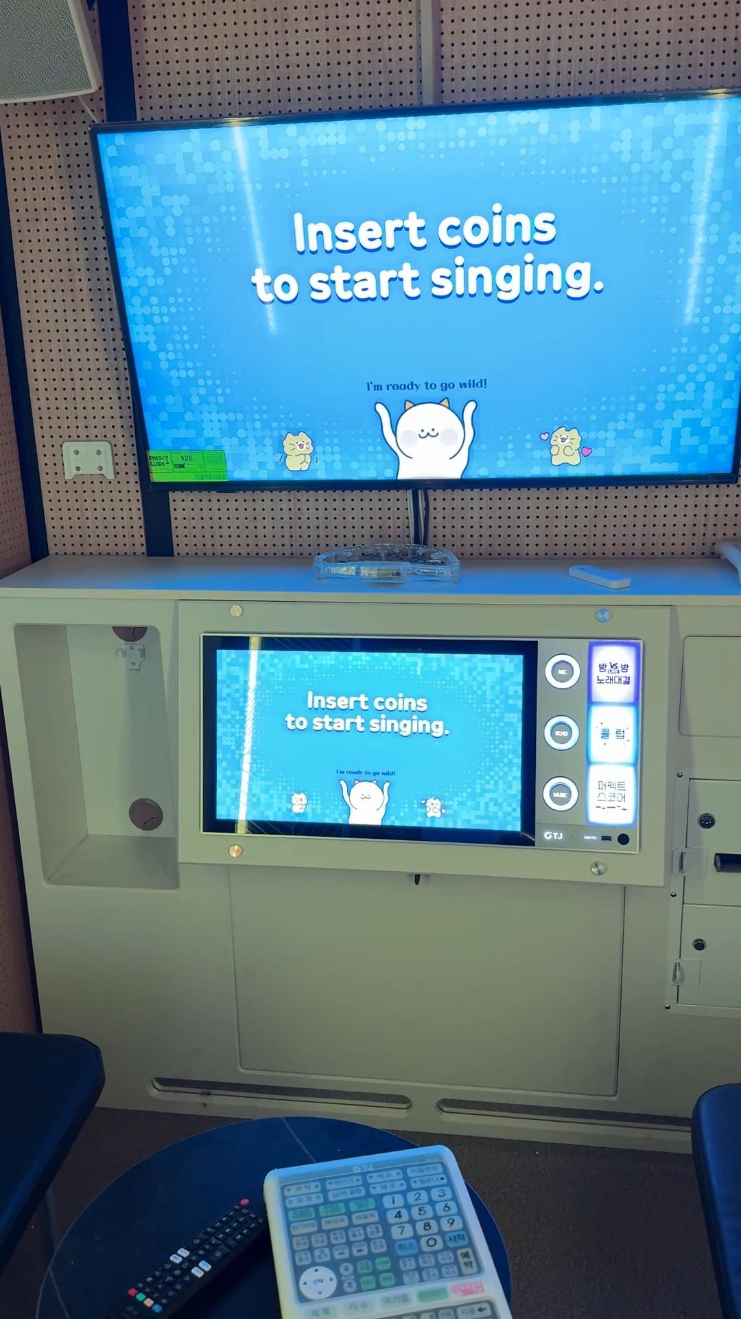 A private karaoke room: a wall-mounted screen reading “Insert coins to start singing” above a TJ Media console, with the song remote and controller on the table.